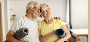 An older man and woman hugging and touching heads together, carrying rolled up yoga mats and smiling. Glad for a better outcome for leg cramp prevention.