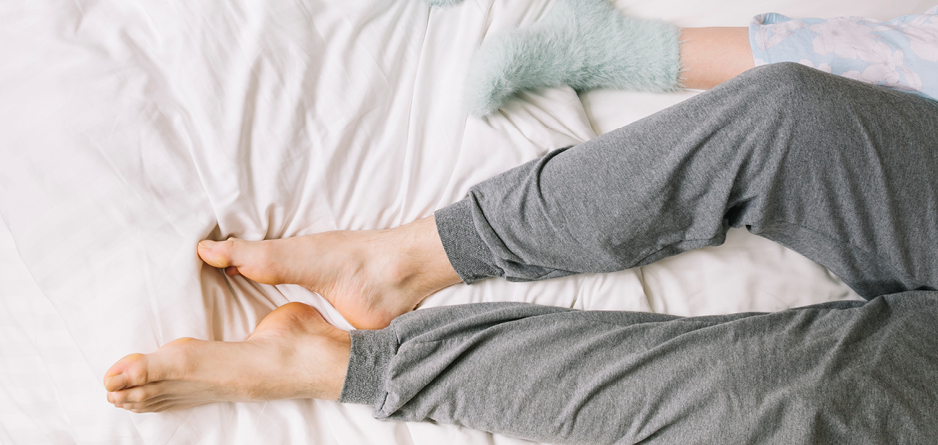 Night time leg cramps causes and solutions two people's legs in a bed, a man with grey sweat pants on and bare feet.